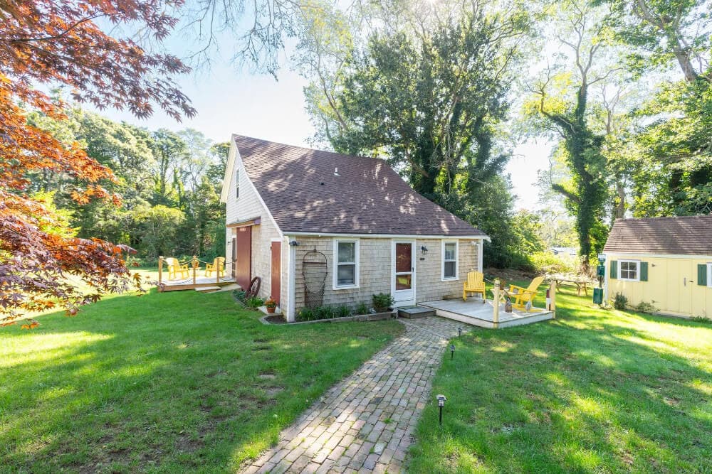 A quaint, two-story house surrounded by greenery, featuring a brick pathway and patio furniture.