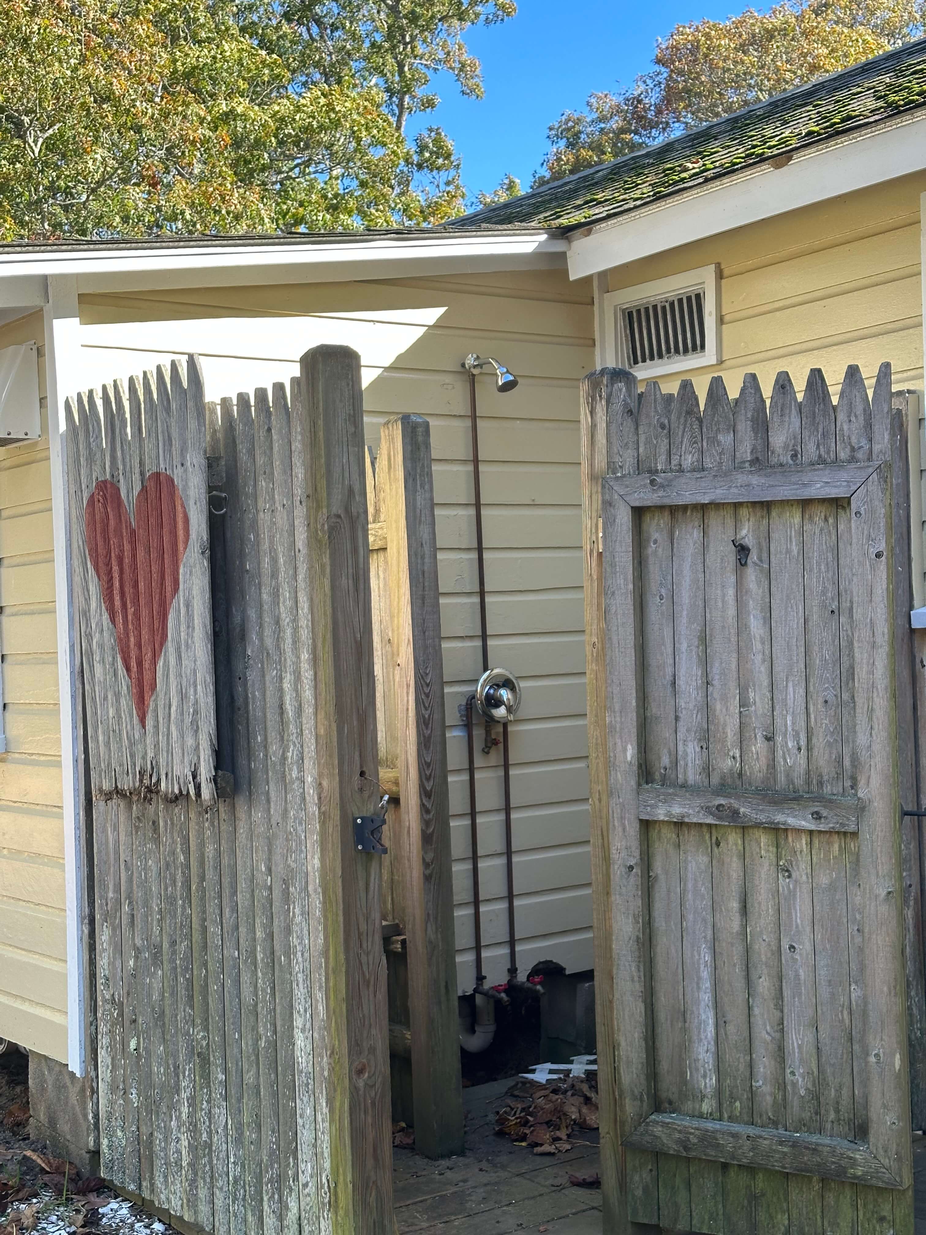 A wooden gate with a red heart leads to a backyard shower area beside a small house.