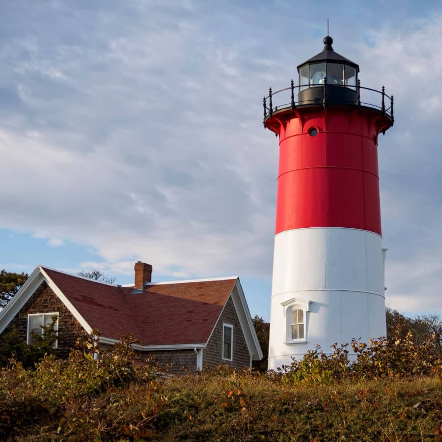 A red and white lighthouse stands beside a small house against a cloudy sky.