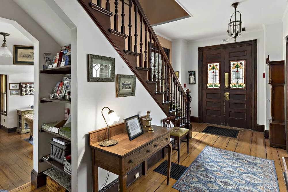 A cozy entryway features a wooden staircase, a small desk with a lamp, and stained glass doors.