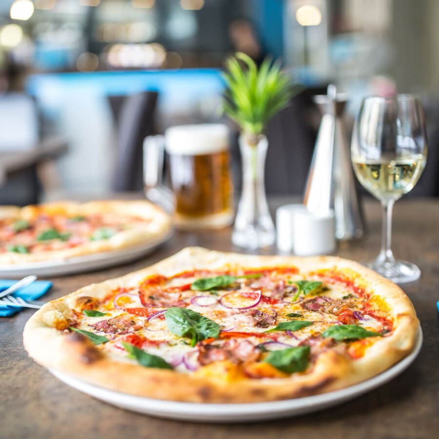 A close-up of a pizza topped with vegetables, served alongside a glass of white wine and a pint of beer in a restaurant setting.
