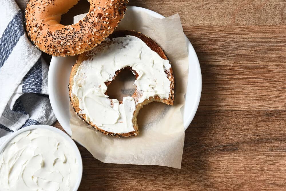 A bagel with cream cheese sits on a plate next to a bowl of cream cheese and a striped napkin.