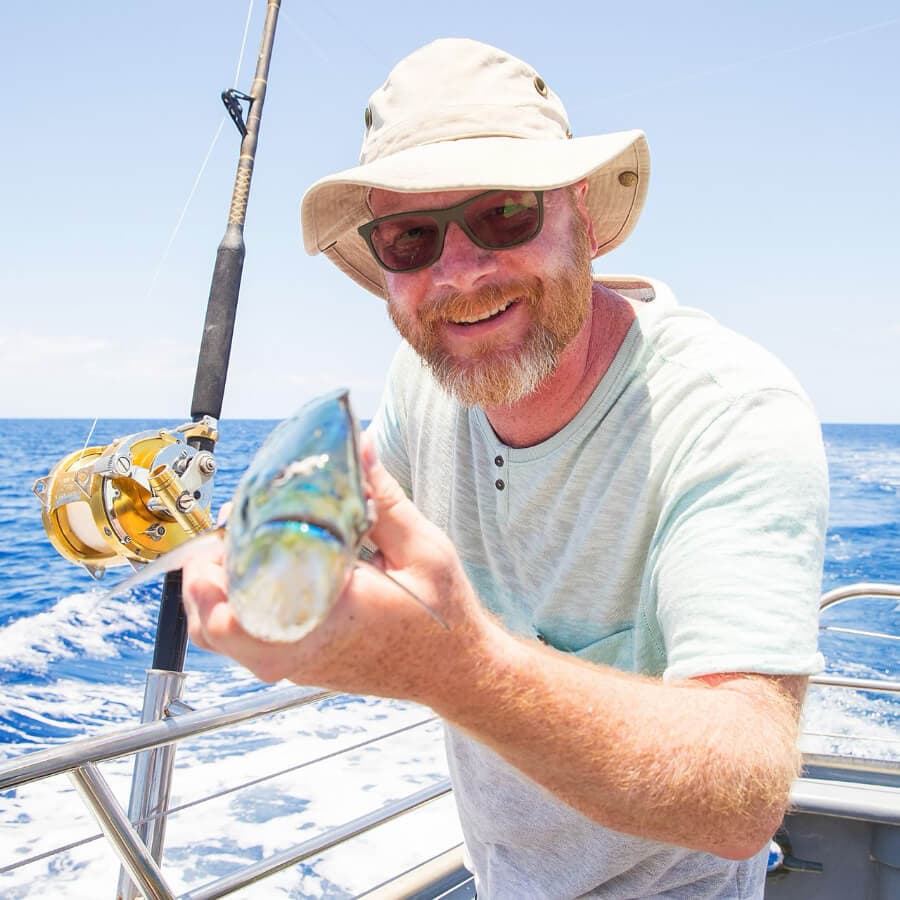 A smiling man in a hat holds up a fish on a boat at sea.