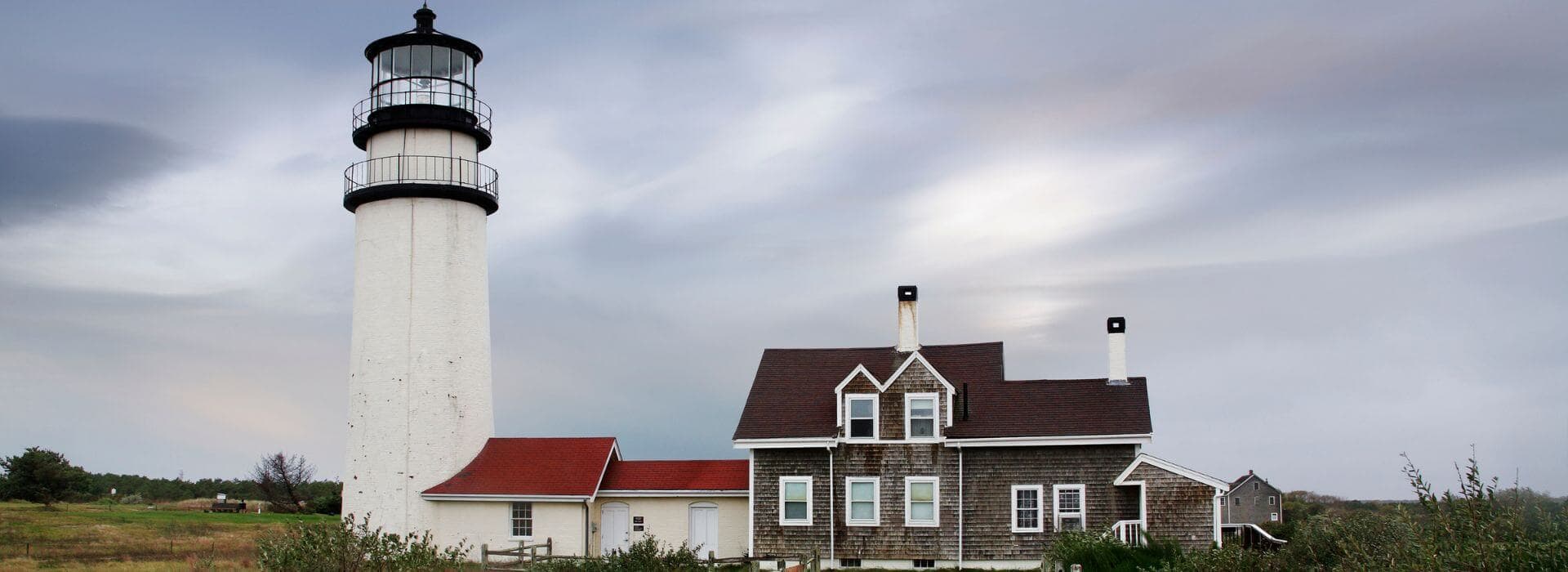 A white lighthouse stands beside a wooden house with a red roof under a cloudy sky.