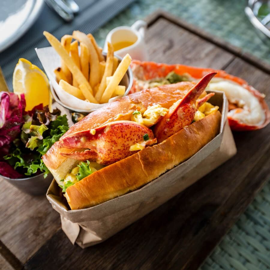 A lobster roll with fries, salad, and lemon on a wooden serving board.