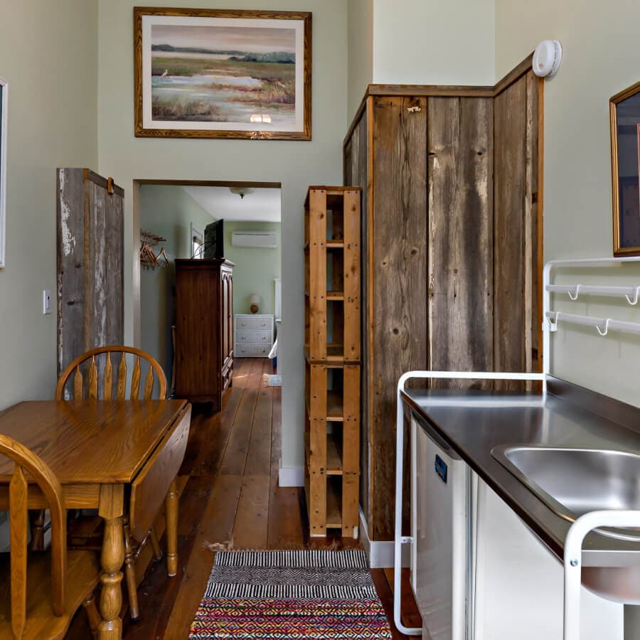 A rustic kitchen area featuring a wooden dining table, a stainless steel sink, and open access to a hallway with wooden furnishings.