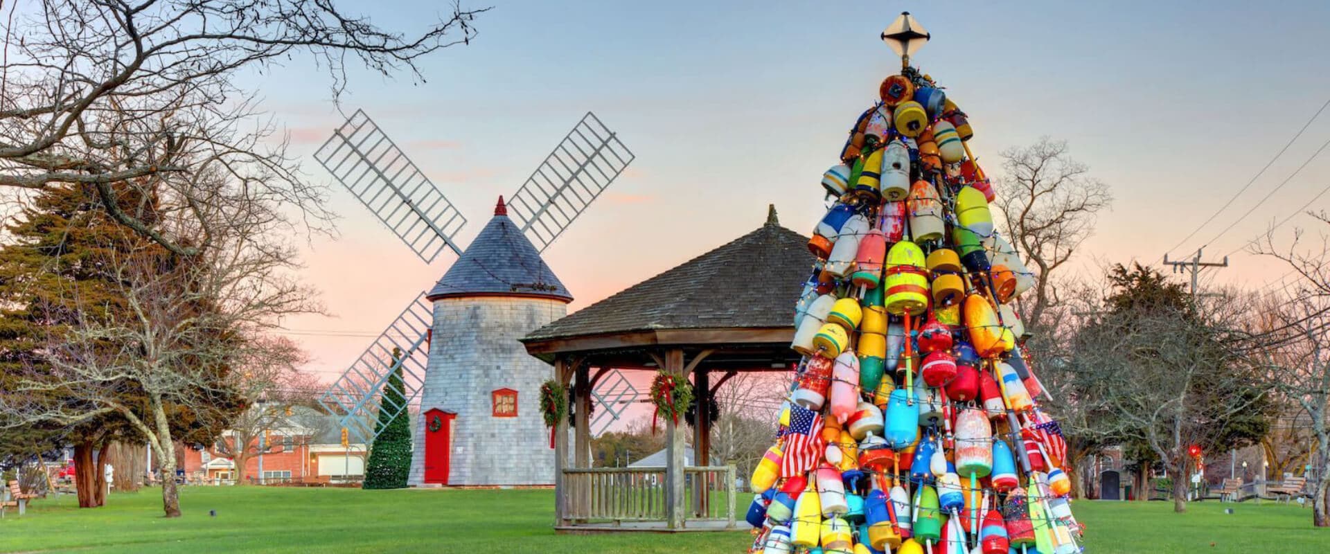 A colorful tower of buoys stands in a park next to a windmill and gazebo against a sunset sky.