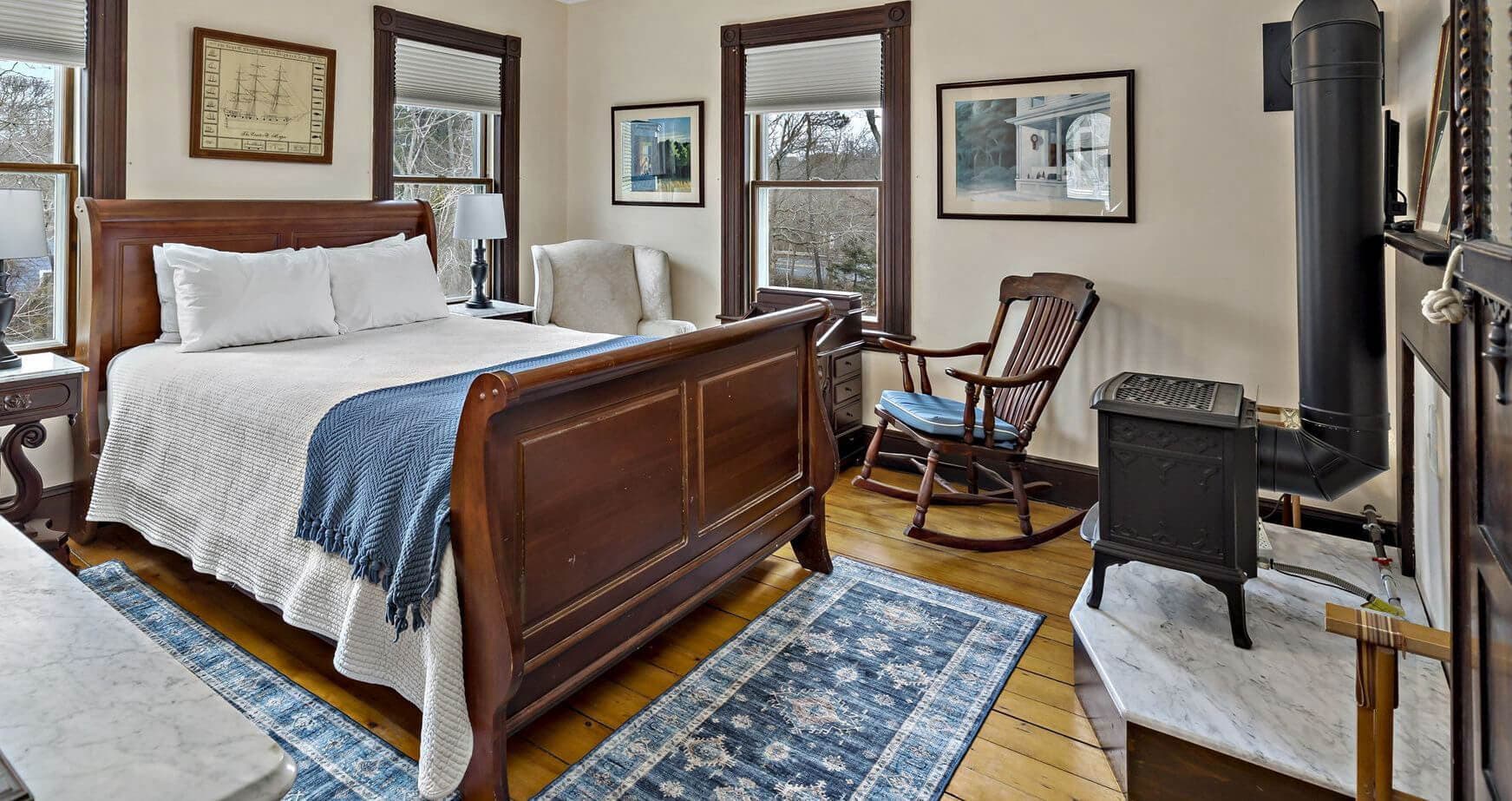 A cozy bedroom featuring a wooden bed, armchair, and stove, with windows showcasing natural light.