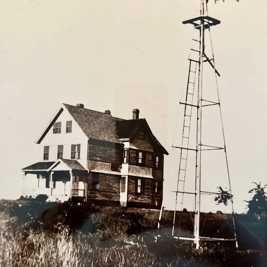 A vintage photo of a two-story wooden house beside a tall windmill.