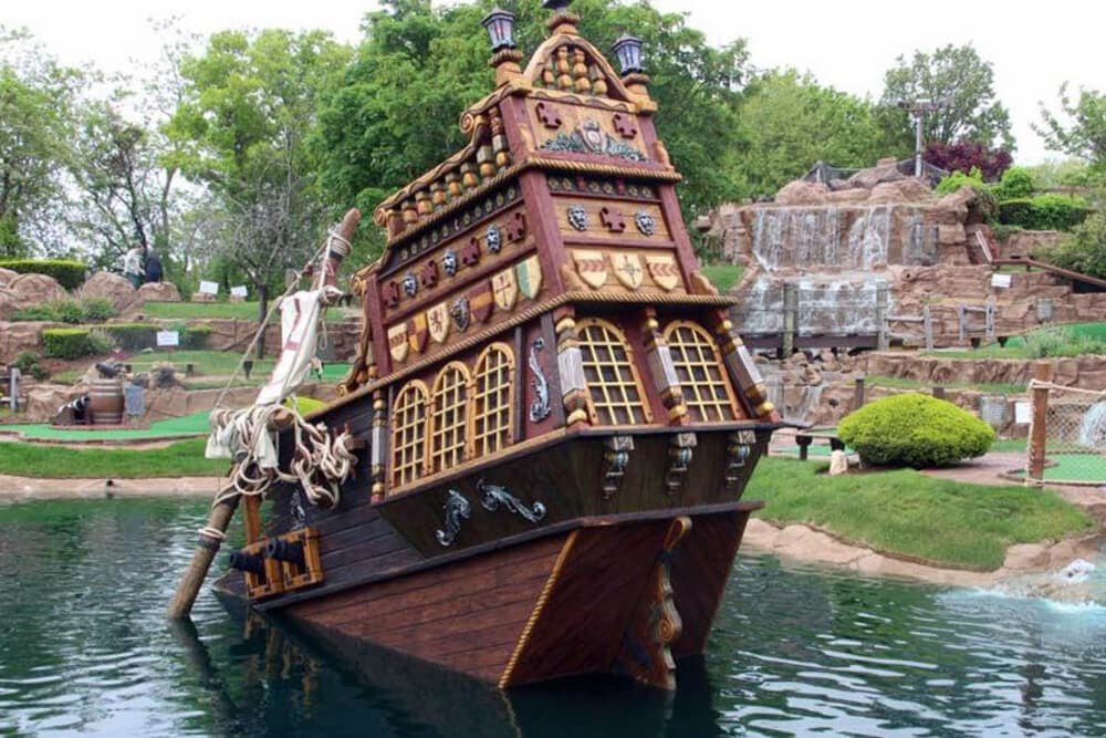 A whimsical pirate ship sculpture partially submerged in a serene pond surrounded by greenery.