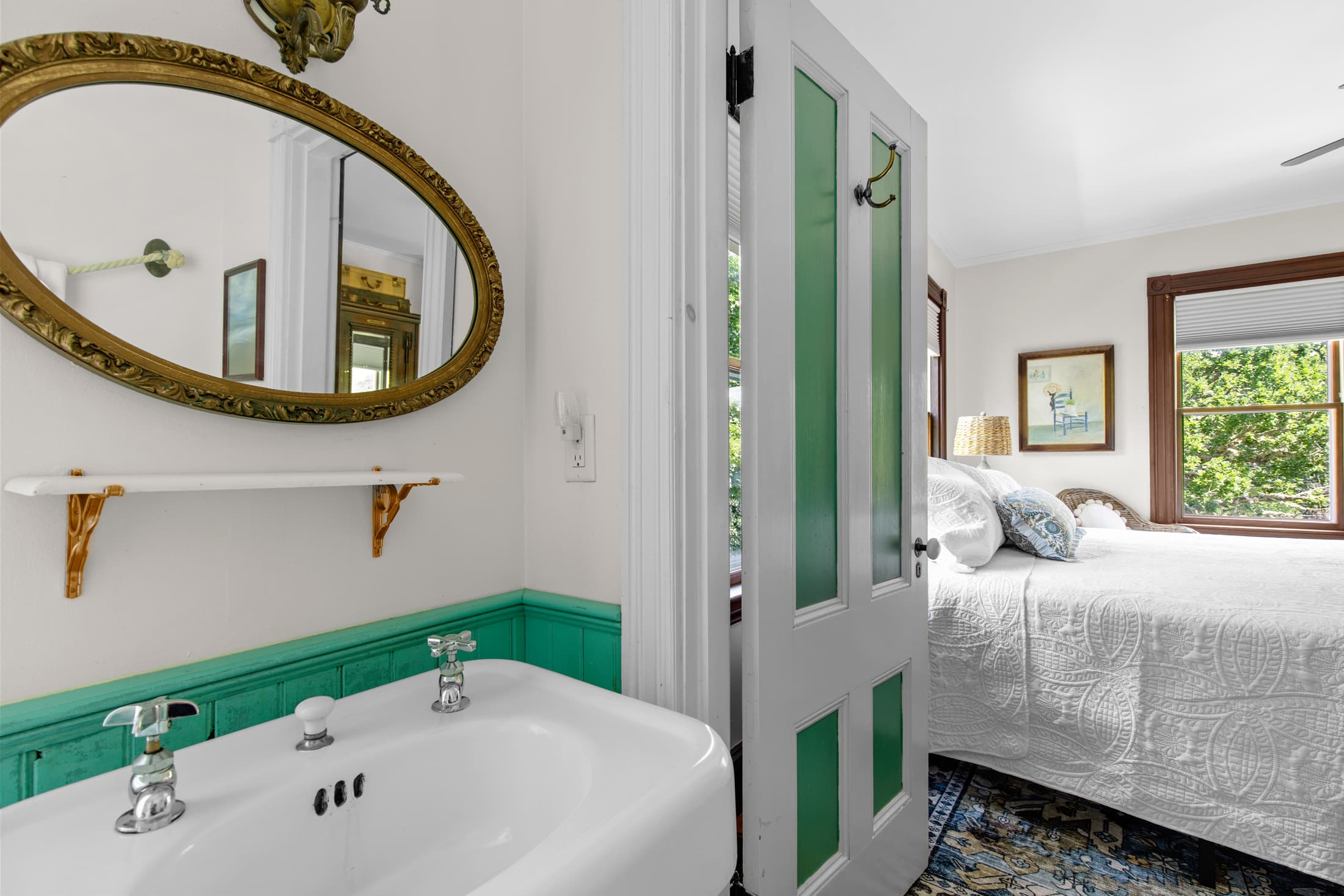 A bright bathroom with a green-bordered sink and mirror, leading to a cozy bedroom with a large window.
