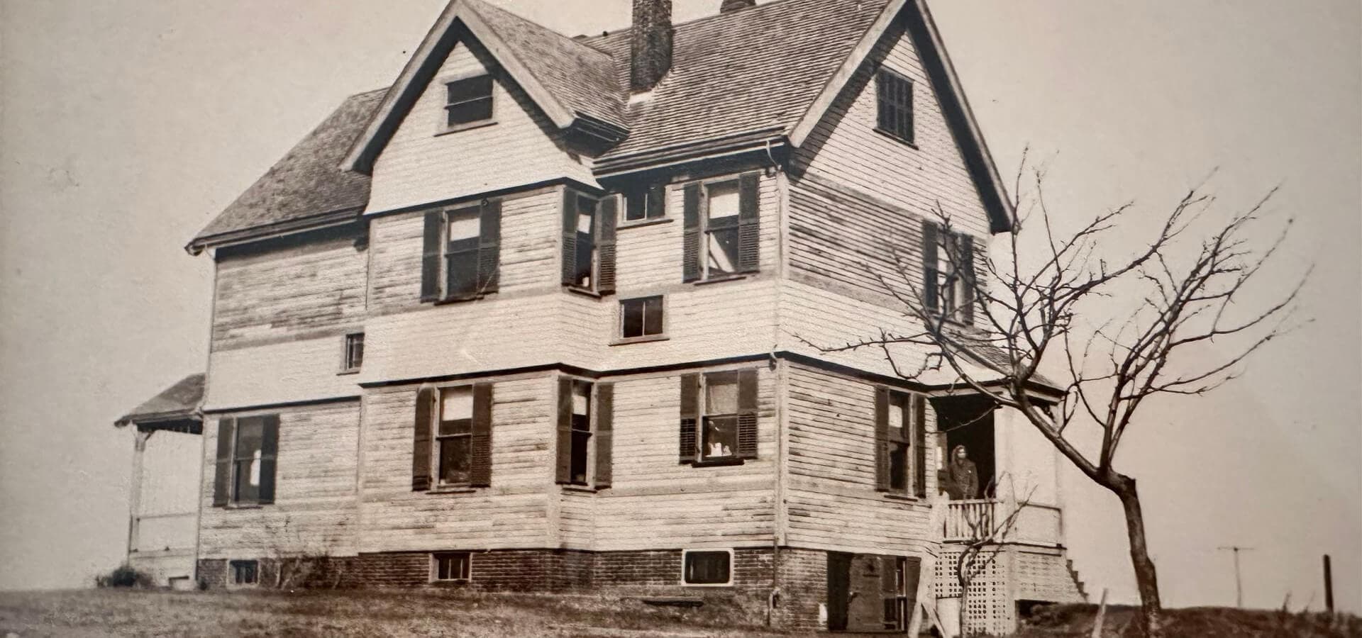 A vintage photograph of a two-story wooden house with multiple windows and a bare tree in the foreground.