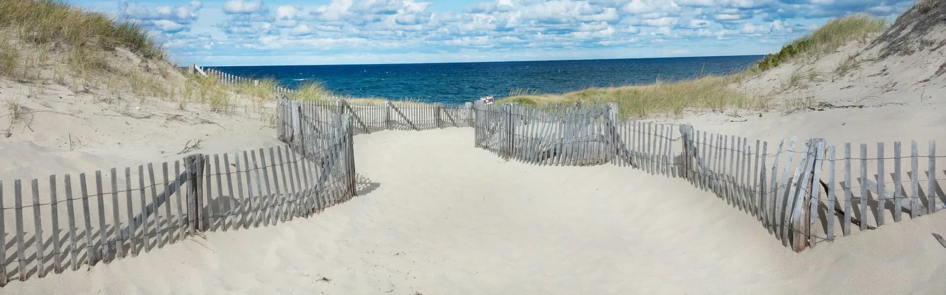 A sandy pathway between grass-covered dunes leads to a calm blue ocean under a partly cloudy sky.