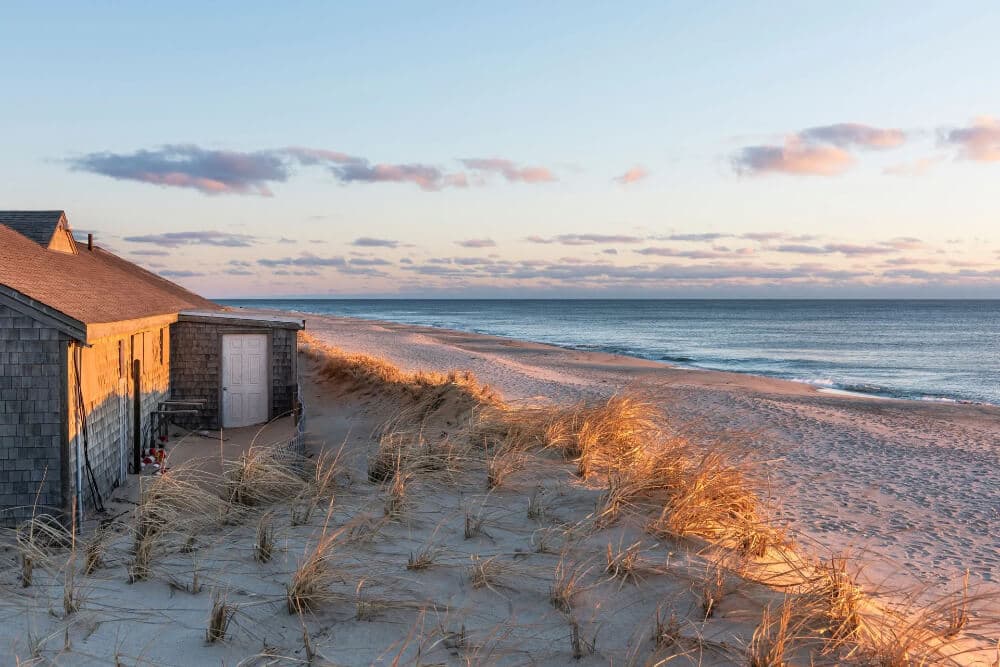 A beachside house sits beside sandy dunes under a colorful sunset sky.