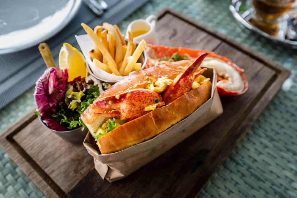 A lobster roll served with fries, salad, and lemon on a wooden platter.