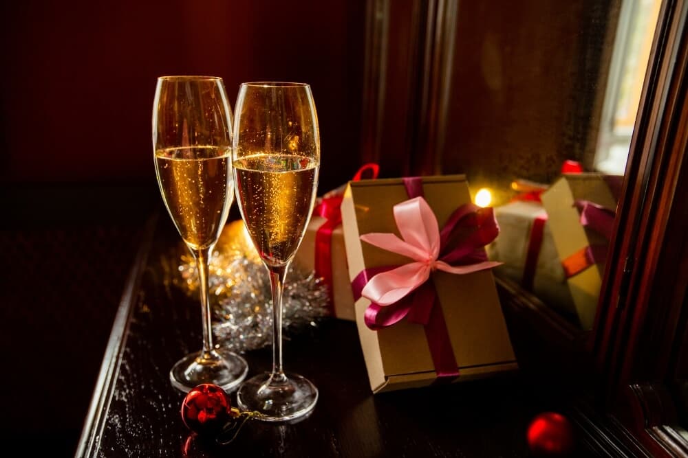 Two champagne flutes beside a beautifully wrapped gift and festive decorations on a reflective surface.