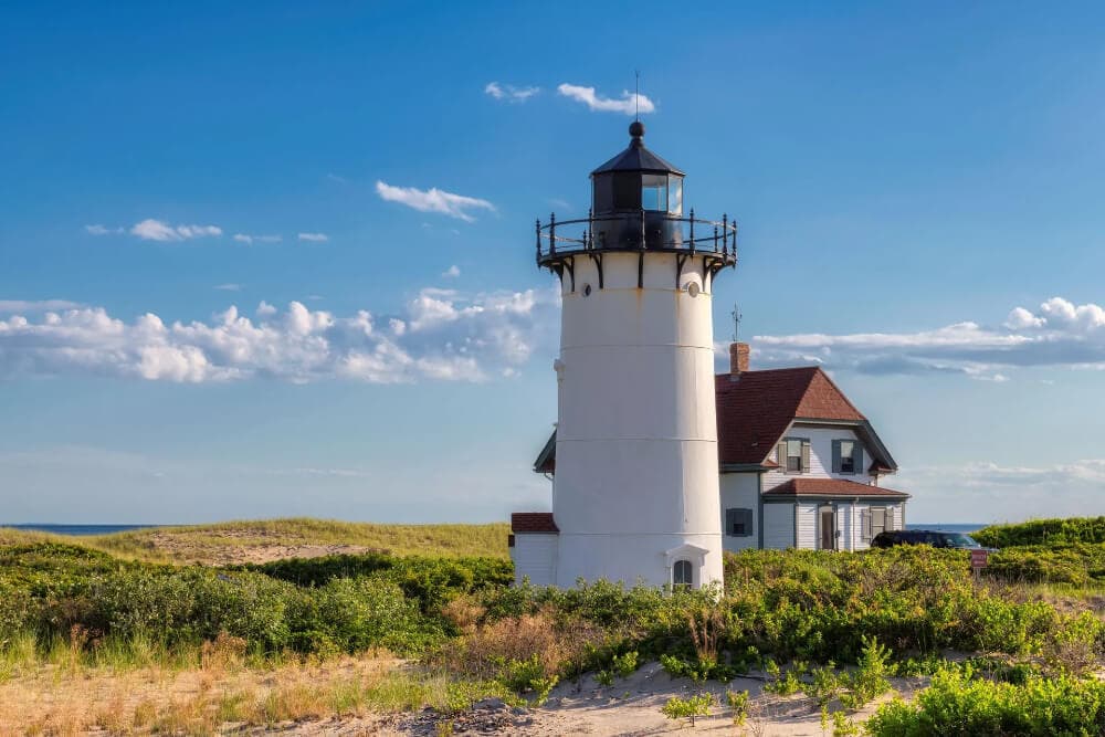 A white lighthouse beside a house, surrounded by green shrubbery and blue sky.