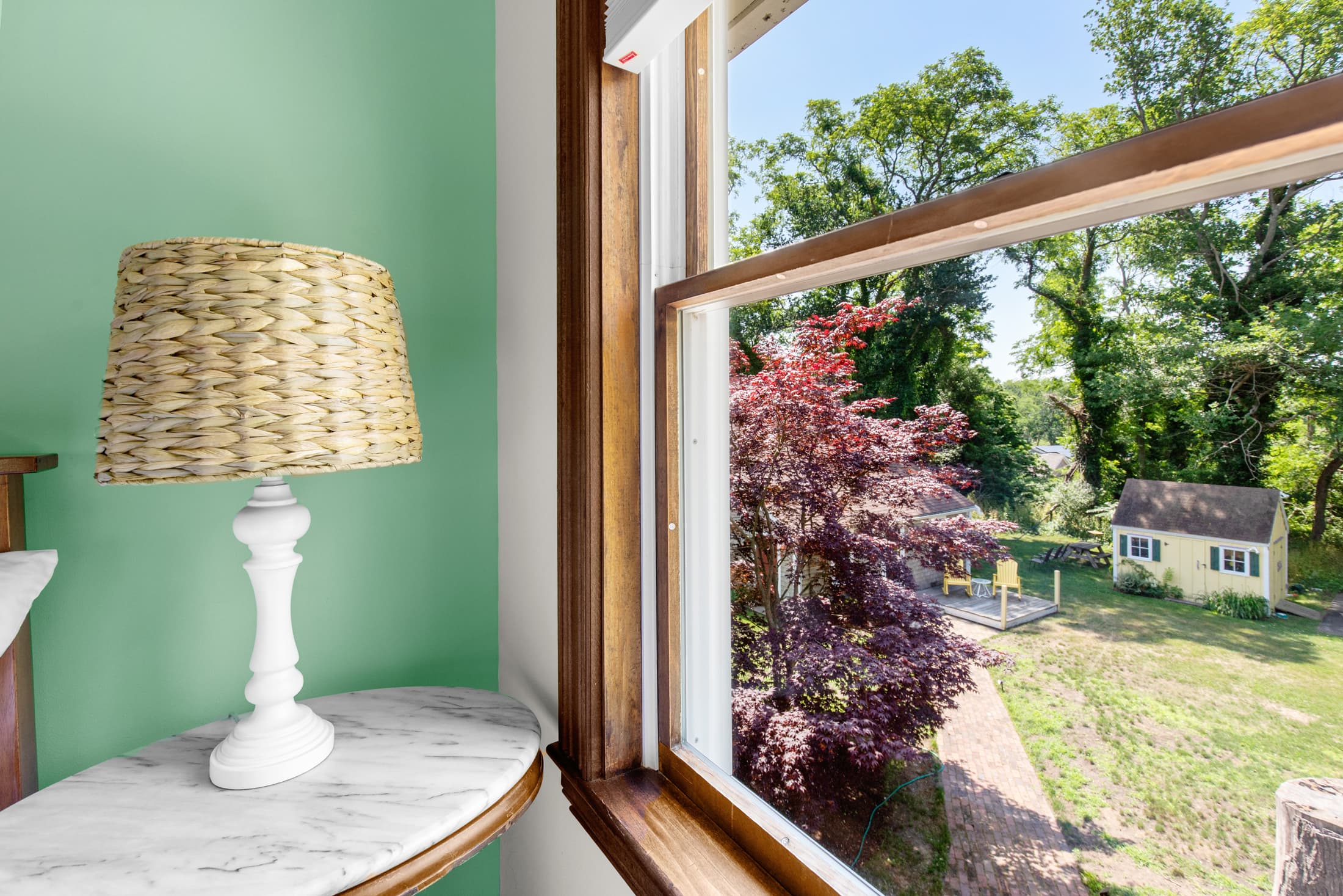A white lamp and marble table beside a window overlooking a garden with trees and a small shed.