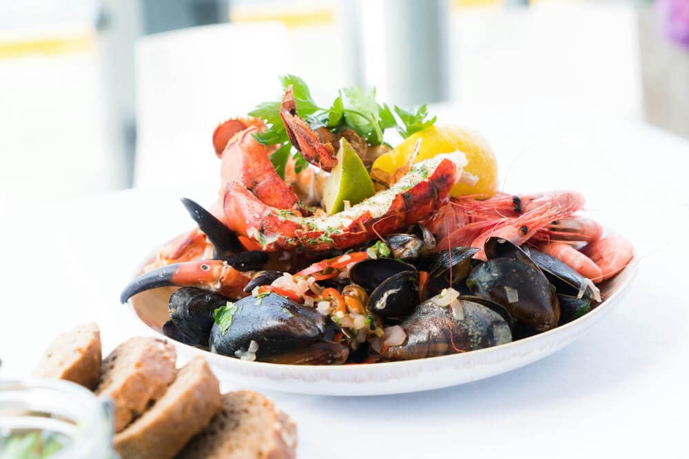 A platter of assorted seafood, including shrimp, lobster, mussels, and garnished with lemon and herbs, accompanied by bread rolls.