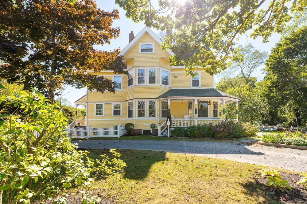 A charming yellow Victorian house surrounded by lush greenery and gardens.