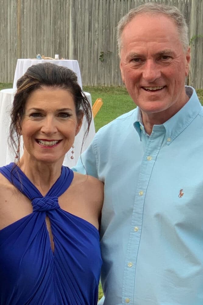 A smiling couple poses together outdoors, with a wooden fence and tables in the background.