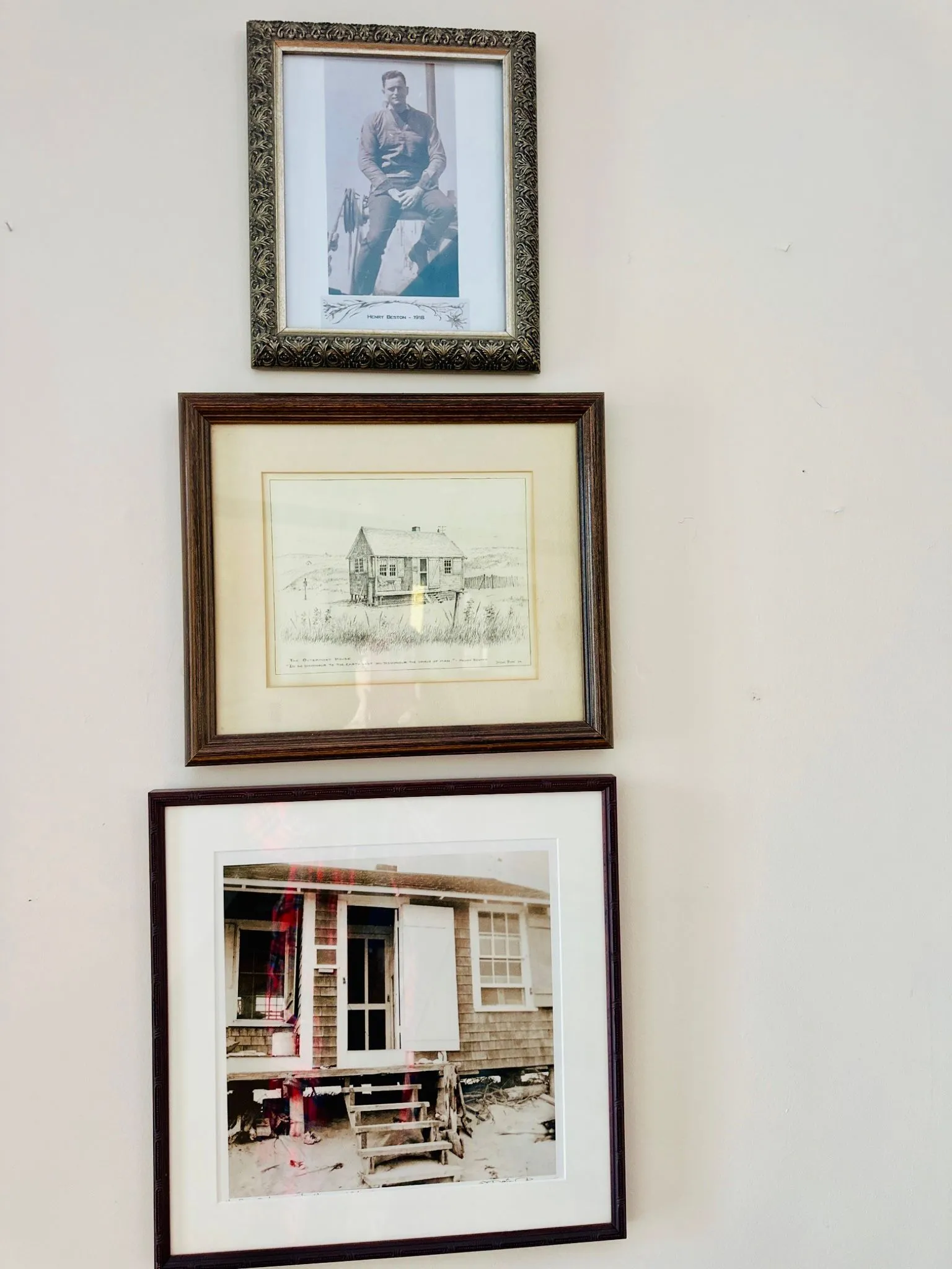 Three framed artworks hang on a wall, including a portrait of a man, a drawing of a house, and a photograph of a weathered building.
