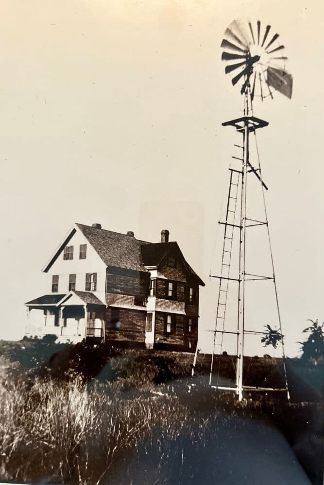A historic farmhouse beside a tall windmill on a grassy landscape.