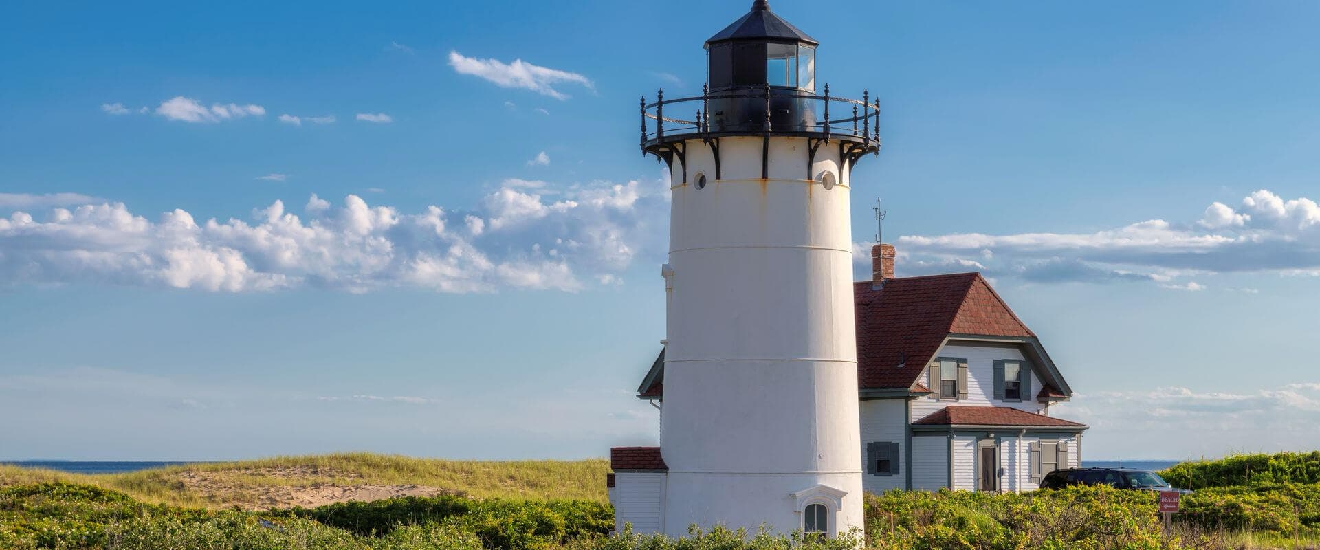 A white lighthouse stands beside a coastal house under a blue sky with scattered clouds.