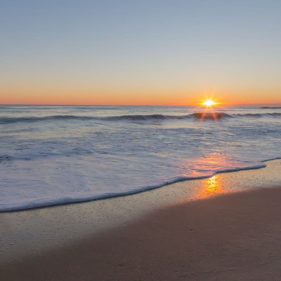 Sunrise over the ocean with gentle waves lapping at a sandy beach.