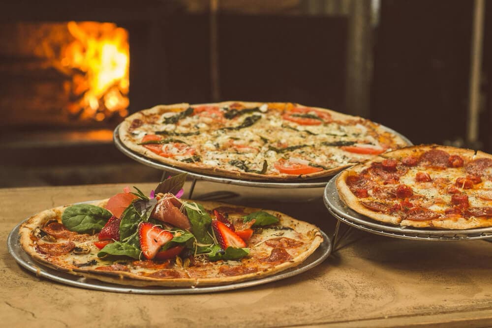 Three pizzas with various toppings in front of a warm fireplace.
