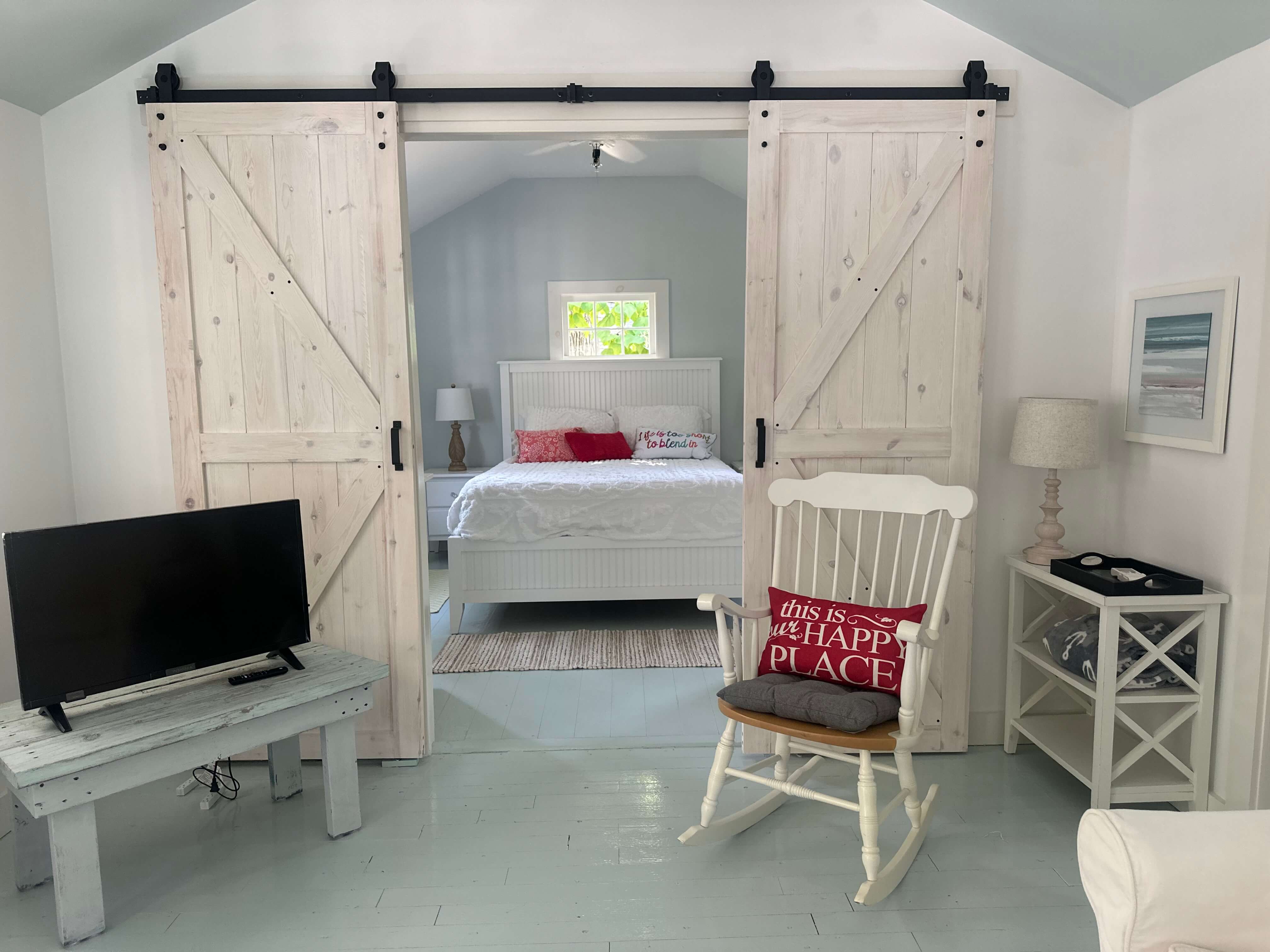 A cozy room featuring barn doors, a white rocking chair, a television, and a bedroom with a bed adorned in red accents.