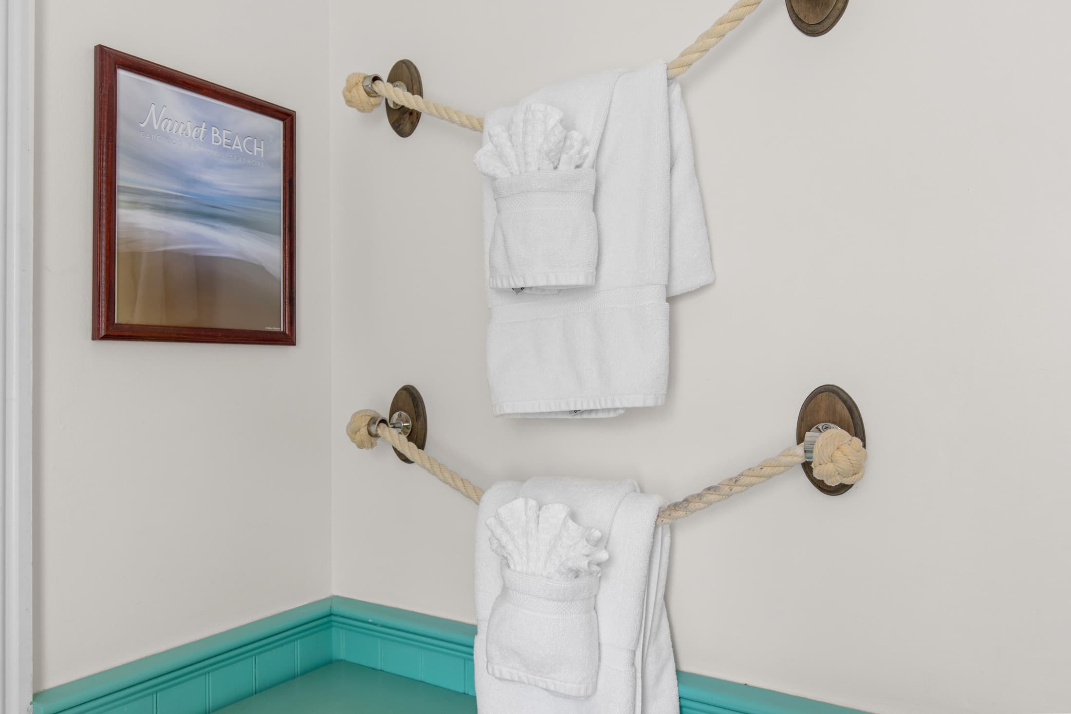 Two neatly hung white towels on a rope rack beside a beach-themed framed picture.