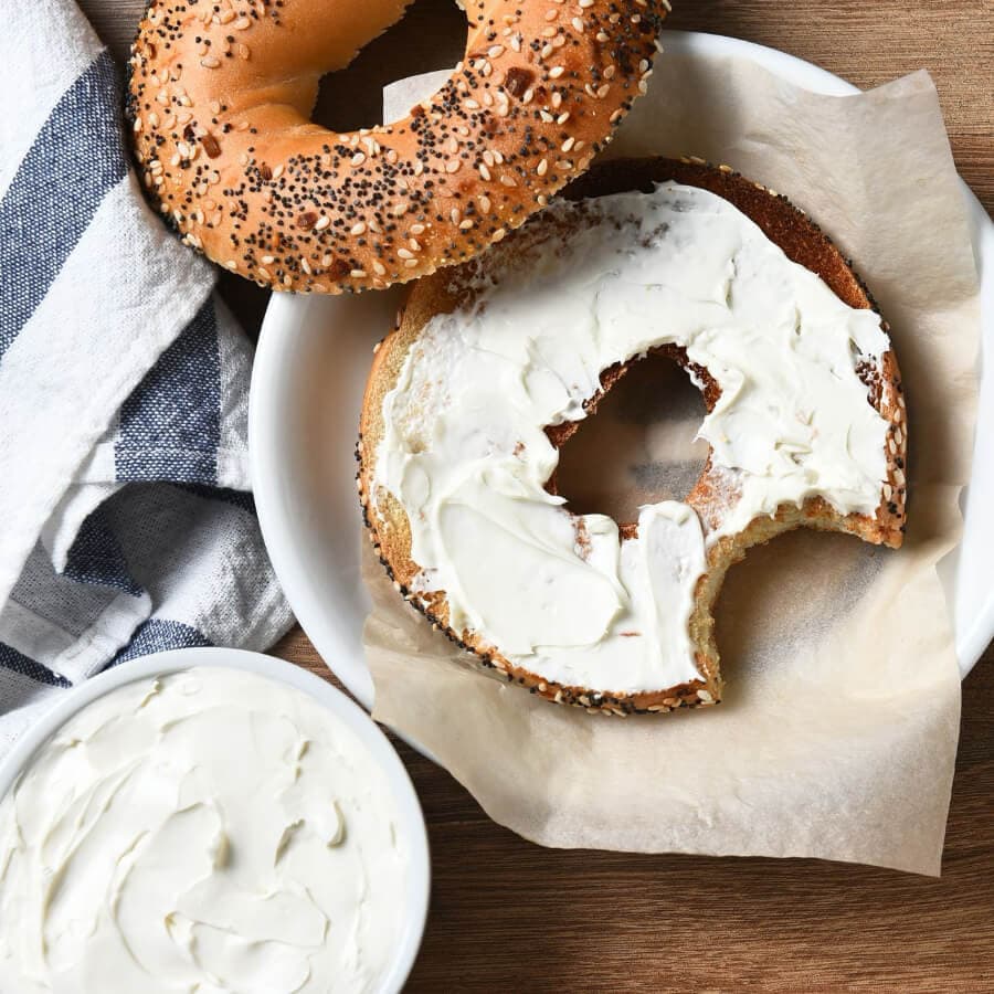 A toasted bagel with cream cheese, a second bagel above, and a bowl of cream cheese on the side.