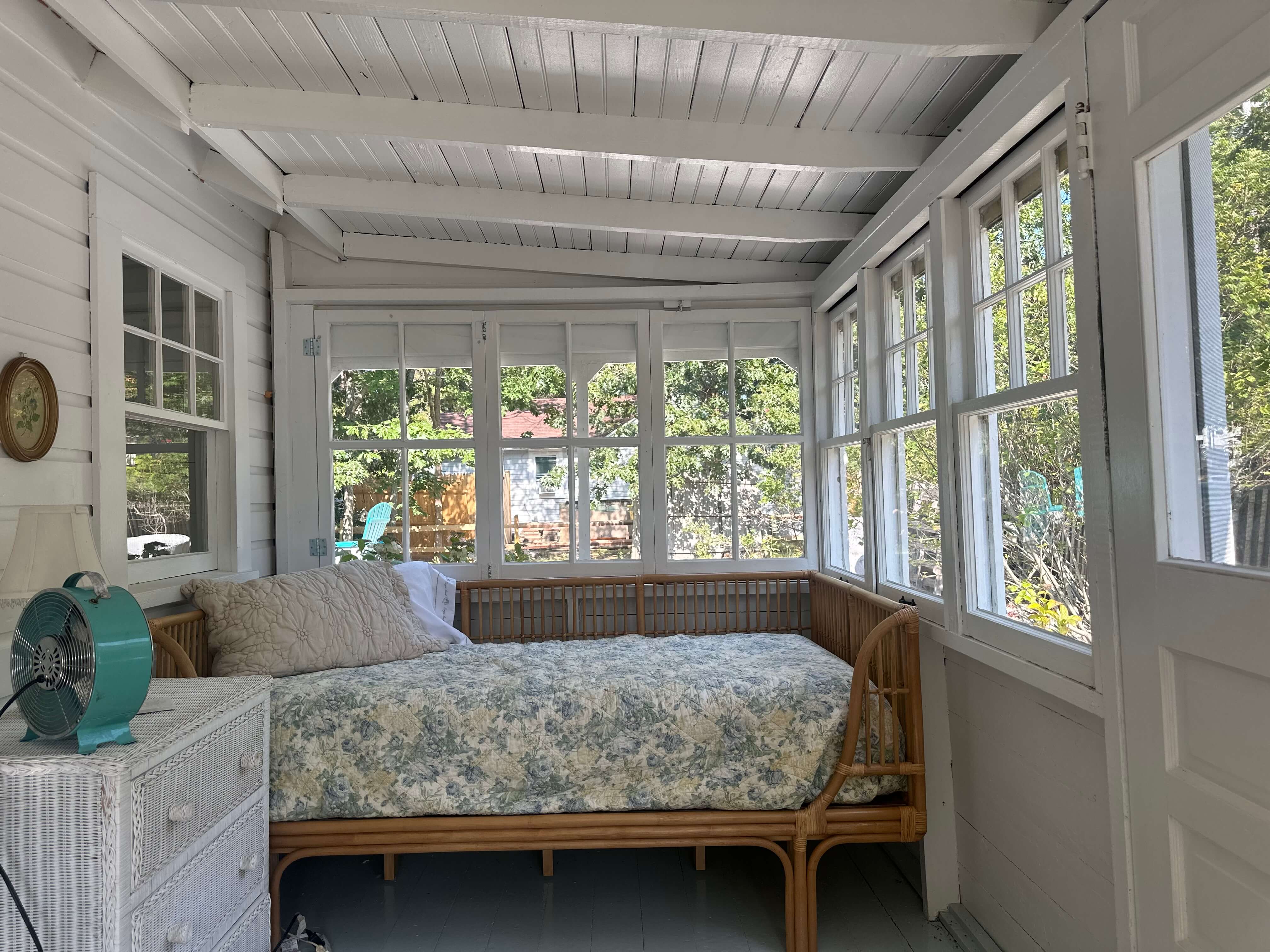 A cozy sunroom featuring a rattan daybed, windowed walls, and a vintage fan.