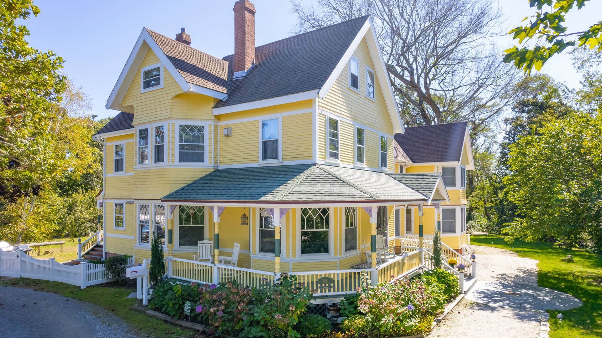A charming two-story yellow Victorian house surrounded by lush greenery.
