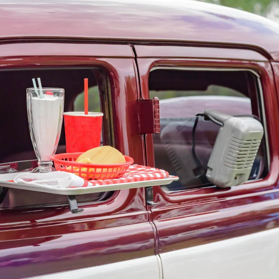A vintage car window displays a tray with a milkshake, soda, and a burger in a red basket.
