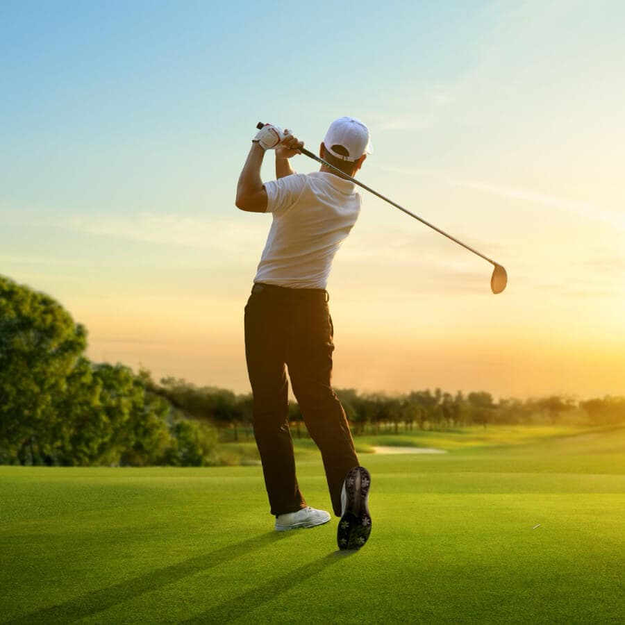 A golfer swings a club at sunset on a lush green course.