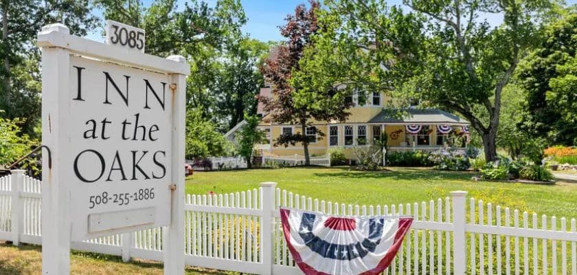 The Inn at the Oaks sign beside a white picket fence, patriotic bunting, green lawn, and yellow inn.